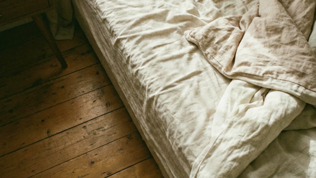 An unmade bed and wooden floor in early morning light